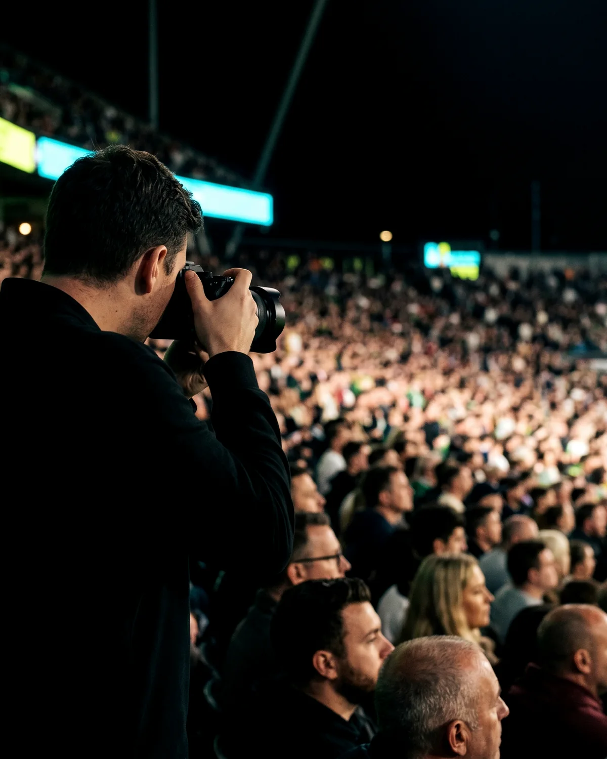 A photographer working the crowd, framing a packed stand at night